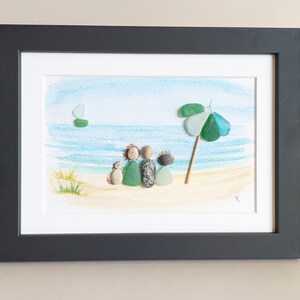 May include: A framed watercolor painting of a beach scene with a family of four and a dog sitting on the sand. The family is made of pebble art, and there is a green and blue umbrella in the background.
