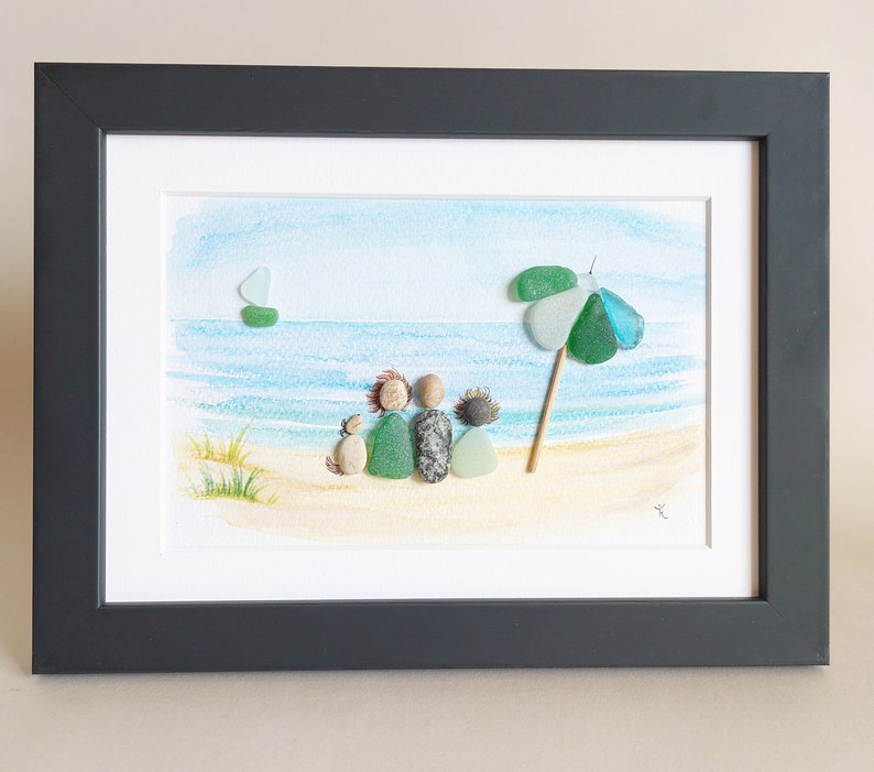 May include: A framed watercolor painting of a beach scene with a family of four and a dog sitting on the sand. The family is made of pebble art, and there is a green and blue umbrella in the background.