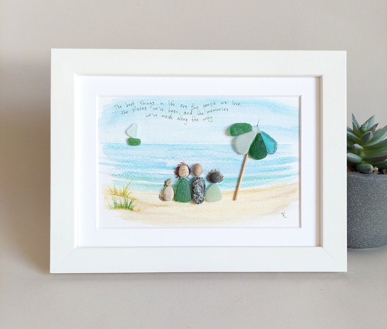 May include: A framed artwork featuring a watercolor painting of a beach scene with a family of four and a dog sitting on the sand. The family is made of sea glass and stones. The quote "The best things in life are the people we love, the places we've been, and the memories we've made along the way" is written in cursive on the white mat.