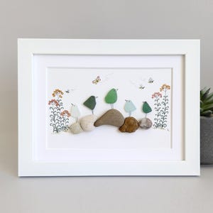 May include: A framed pebble art piece depicting a group of five birds perched on rocks, with green, blue, and white painted details. The birds are surrounded by small flowers and butterflies, creating a whimsical nature scene.