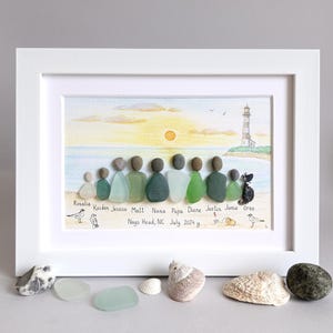 May include: A framed watercolor painting of a beach scene with a lighthouse in the distance.  The painting features a group of sea glass figures representing a family, with names written below each figure.  The text below the figures reads "Nags Head, NC July 2024 y."
