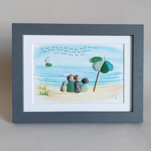 May include: A framed artwork featuring a watercolor painting of a beach scene with a family of four figures made from sea glass and a small sailboat. The quote "The best things in life are the people we love, the places we've been, and the memories we've made along the way" is written in black ink on the white background.