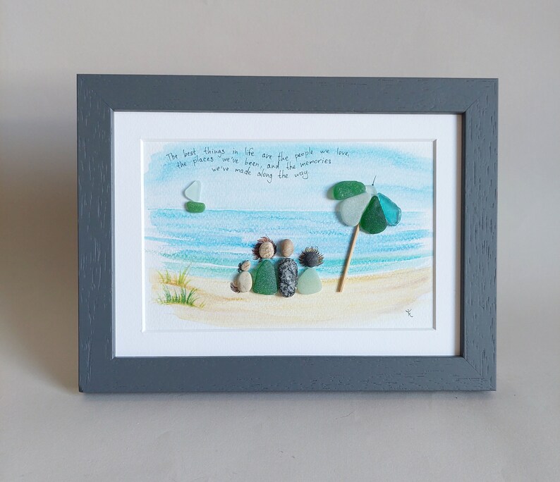 May include: A framed artwork featuring a watercolor painting of a beach scene with a family of four figures made from sea glass and a small sailboat. The quote "The best things in life are the people we love, the places we've been, and the memories we've made along the way" is written in black ink on the white background.