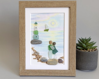 May include: A framed pebble art piece featuring a lighthouse made of sea glass and stones, a small sailboat, and two figures standing on a beach. The background is a watercolour painting of a soft sky and sea with a sun, all in a light wood frame.