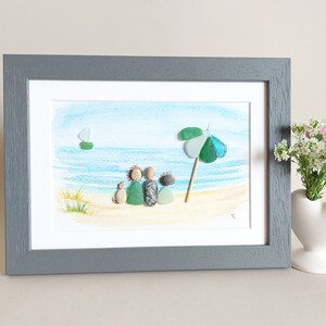 May include: A framed watercolor painting of a beach scene with a family of four and a dog sitting on the sand under a green and blue umbrella. The painting is in a gray frame.
