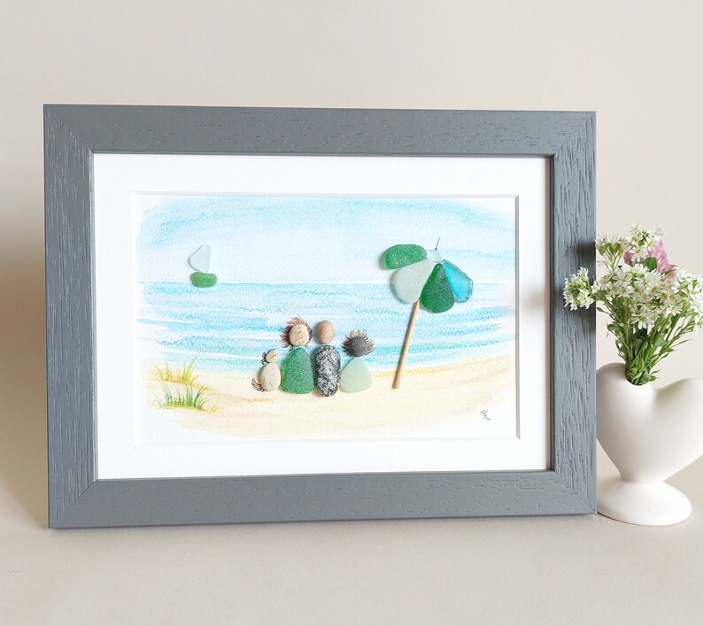 May include: A framed watercolor painting of a beach scene with a family of four and a dog sitting on the sand under a green and blue umbrella. The painting is in a gray frame.