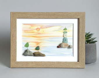 May include: A framed mixed media artwork featuring a watercolour sunset background with a lighthouse and two birds constructed from smooth pebbles and sea glass. The scene includes a light wooden frame and a white mat, depicting a coastal landscape.