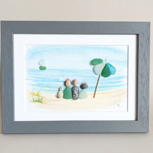May include: A framed watercolor painting of a beach scene with a family of four and a dog sitting on the sand. The family is made of pebble art, and there is a green and blue beach umbrella in the background.