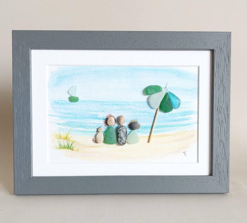 May include: A framed watercolor painting of a beach scene with a family of four and a dog sitting on the sand. The family is made of pebble art, and there is a green and blue beach umbrella in the background.