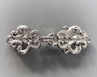 May include: Silver-toned metal clasp with a decorative, ornate design. The clasp is made of two pieces that connect in the middle.