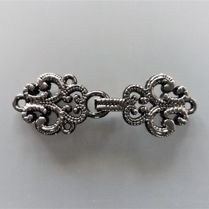 Könnte beinhalten: Silberfarbener Metallverschluss mit einem dekorativen, verzierten Design. Der Verschluss hat zwei Schlaufen auf der einen Seite und eine Verbindungsstange auf der anderen.