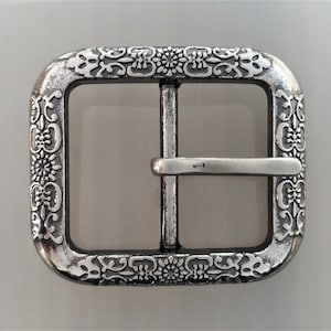 Pode incluir: Fivela de cinto de metal prateado com um design floral ornamentado. A fivela é retangular com cantos arredondados e um acabamento liso e polido.