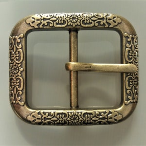 May include: A gold-toned metal belt buckle with an ornate, engraved design. The buckle is rectangular with rounded corners and a centre bar for attaching a belt.