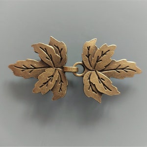 Può includere: Un paio di chiusure a forma di foglia in metallo color oro antico con un anello decorativo al centro. Le chiusure sono progettate per essere utilizzate come chiusura decorativa per abbigliamento o accessori.