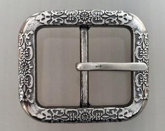 May include: Silver-toned metal belt buckle with an ornate floral design. The buckle has a rectangular shape with rounded corners and a smooth, polished finish.