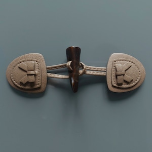 Peut inclure: Une fermeture à bascule beige avec un bouton en forme de corne marron foncé. La fermeture comprend deux pièces en cuir cousues et arrondies avec un motif décoratif, reliées par un cordon en cuir beige. Le fond est gris uni.