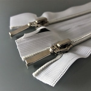 May include: Two white zipper tapes with silver metal sliders. The zippers are laid out flat on a gray surface.