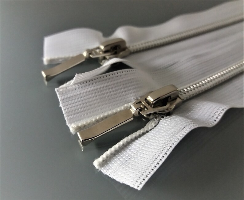 May include: Two white zipper tapes with silver metal sliders. The zippers are laid out flat on a gray surface.