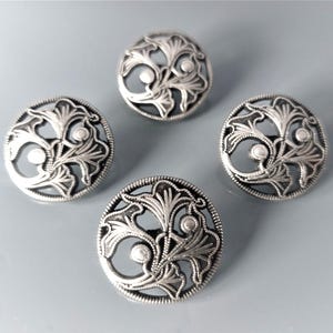 Può includere: Quattro bottoni metallici circolari color argento con un design traforato di motivi floreali o a foglia stilizzati. Ogni bottone presenta un motivo in rilievo e testurizzato con intagli complessi e piccoli accenti circolari.