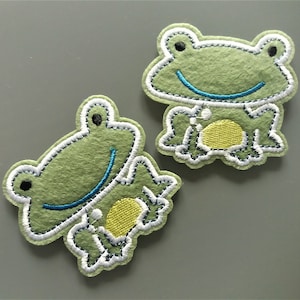 Peut inclure: Deux appliques de grenouille en feutre vert avec des détails brodés blancs et bleus. Les grenouilles ont des ventres brodés jaunes et sourient.