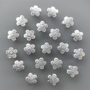 May include: A collection of 20 silver flower-shaped charms. The charms are all the same size and shape, and they have a slightly raised design.