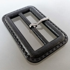 May include: A black plastic belt buckle with a silver metal slider. The buckle has a textured surface and a decorative border.