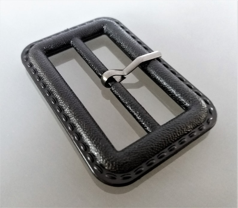May include: A black plastic belt buckle with a silver metal slider. The buckle has a textured surface and a decorative border.