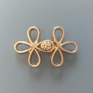 Könnte beinhalten: Goldfarbener Metallverschluss mit einem dekorativen Knotendesign. Der Verschluss besteht aus zwei ineinandergreifenden Teilen, die jeweils die Form eines Kleeblatts haben.