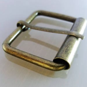 May include: An antique brass-finished metal buckle with a roller bar. The rectangular buckle features a square frame with rounded corners and a central bar. The metal has a slightly aged, worn appearance, suggesting vintage or rustic style.