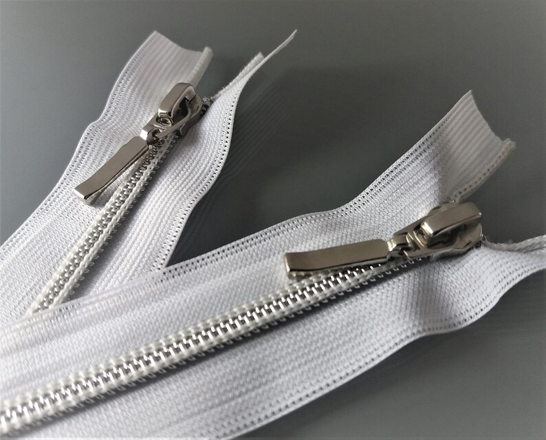 May include: Two white zipper pulls with silver metal sliders. The zippers are open and the sliders are in the middle of the zipper tape.
