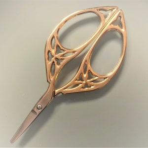 Könnte beinhalten: Goldfarbene dekorative Schere mit einem komplizierten floralen Design. Die Schere hat eine spitze Spitze und eine kleine Schraube in der Mitte.