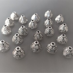 May include: A collection of 20 silver-toned metal cone-shaped beads with an ornate design. The beads have a hole in the top for stringing.