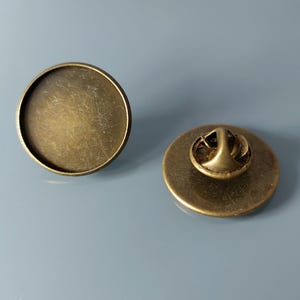 Op de afbeelding: Twee ronde reversspelden in antiek messingkleur. Eén speld heeft een plat, cirkelvormig oppervlak met een verzonken gedeelte. De andere speld toont de achterkant met een speldsluiting. De spelden liggen op een lichtblauwe ondergrond.
