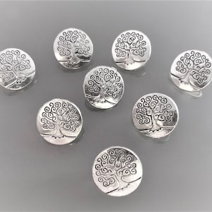 Op de afbeelding: Een verzameling zilverkleurige, ronde metalen knopen. Elke knoop heeft een gedetailleerd boomontwerp met wervelende takken en een getextureerde afwerking. De knopen liggen op een grijze ondergrond, waardoor hun ingewikkelde details worden benadrukt.