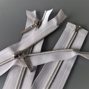 May include: Three white zipper tapes with silver metal sliders. The zippers are laid out on a gray surface.
