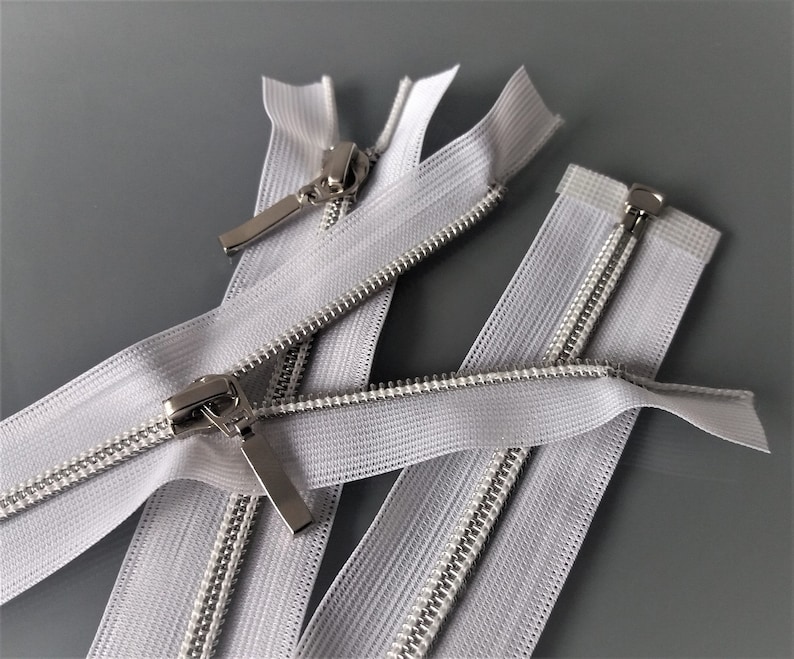 May include: Three white zipper tapes with silver metal sliders. The zippers are laid out on a gray surface.