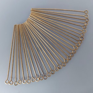 Peut inclure: Collection d'épingles à œil de couleur laiton disposées en éventail. Chaque épingle a un corps en fil droit et une extrémité bouclée. Les épingles sont présentées sur un fond gris clair.