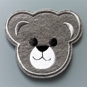 Peut inclure: Applique en feutre gris d'un visage d'ours en peluche avec des détails brodés blancs et noirs.
