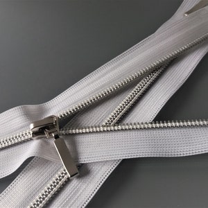 May include: White zipper tape with a silver metal slider. The zipper is open and the tape is folded over itself.