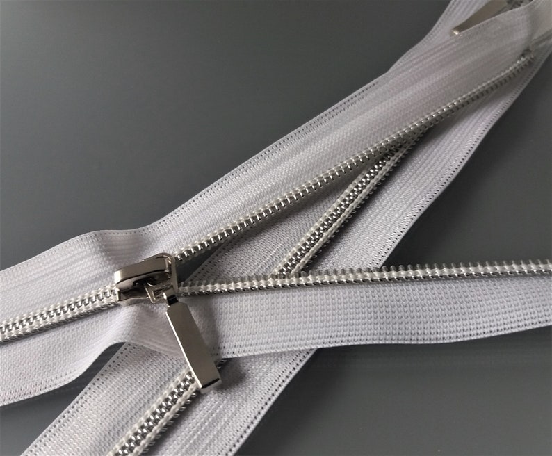 May include: White zipper tape with a silver metal slider. The zipper is open and the tape is folded over itself.