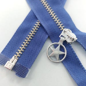 May include: A close-up shot of a blue zipper with silver metal teeth and a star-shaped pull. The zipper is made of a strong fabric and is suitable for clothing or sewing projects. The star pull adds a decorative touch.