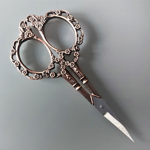 Pode incluir: Um par de tesouras ornamentadas de estilo antigo com um design floral. As tesouras são feitas de metal e têm um acabamento bronze. As lâminas são afiadas e pontiagudas.