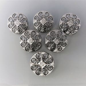 May include: Six silver-toned buttons with a decorative, intricate, filigree design. Each button features a diamond-shaped centre with a swirl pattern.