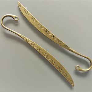 May include: Two gold-toned metal bookmarks with decorative swirls. One bookmark has a hook at the end, the other has a pointed tip.