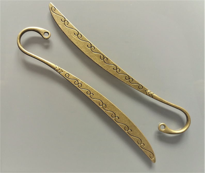 May include: Two gold-toned metal bookmarks with decorative swirls. One bookmark has a hook at the end, the other has a pointed tip.