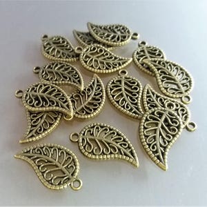 Op de afbeelding: Een verzameling van 14 goudkleurige metalen bladeren met ingewikkelde filigraanontwerpen. Elke hanger heeft een lus om aan sieraden of andere knutselprojecten te bevestigen.