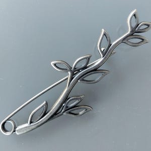 Peut inclure: Broche argentée en forme de branche feuillue. La broche est dotée d'un fermoir de type épingle de sûreté et est ornée de feuilles ajourées délicates. Le métal présente un aspect légèrement vieilli, lui conférant un aspect vintage.