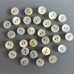 May include: A collection of 27 white oval-shaped buttons with two holes each. The buttons are made of mother of pearl and have a subtle iridescent sheen.