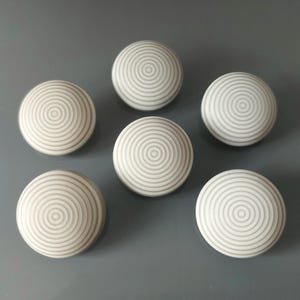 Może przedstawiać: Sześć białych gałek do szafek z wzorem koncentrycznych okręgów. Gałki są ułożone na szarej powierzchni. Gałki są okrągłe i mają lekko wypukłą, teksturowaną powierzchnię.