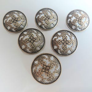 Pode incluir: Seis botões de metal dourado antigo com um design floral decorativo. Os botões têm forma circular com um centro elevado e uma borda festoneada.